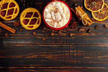 A cup of hot drink with whipped cream, marshmallows and powder, in a knitted cover and homemade cookies, cinnamon, Christmas decoration, lie on a wooden table. Copy space.