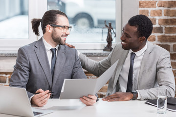 smiling multicultural business partners sitting at table looking at each other in office