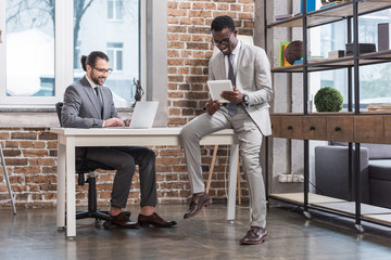 handsome man typing on laptop keyboard and african american business partner sitting on table with digital tablet