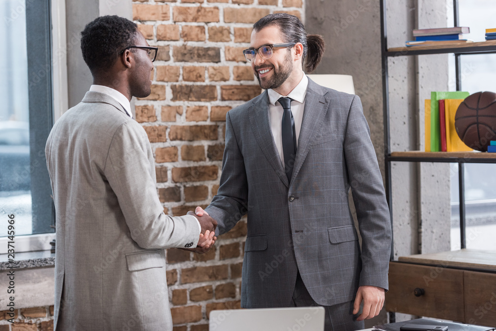 Wall mural smiling handsome businessman and african american partner shaking hands in office