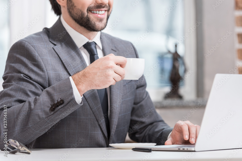 Wall mural cropped view of smiling businessman drinking coffee and typing on laptop keyboard at table