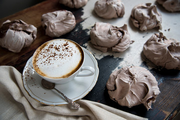 A cup of aromatic coffee with milk and cinnamon on a rustic table, next to the chocolate meringue and an old spoon and linen cloth