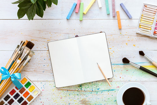 Colorful creative table with blank notebook for sketches and paints, pencil, paintbrushes set and cup of coffee on white wooden table, top view, copy space, flat lay style, drawing class education