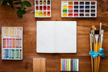 Creative art work accessory supplies set, open notebook for sketch, paint brushes, paintbox with watercolors, crayons, pencils on brown wooden artistic background, back to school top view copy space