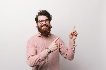 Portrait of happy cheerful man with christmas decorated beard pointing at copyspace over white...