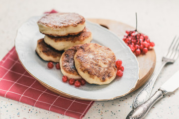 On a white table made of artificial stone is a plate with five cottage cheese pancakes and red berries . On the table is wooden cutting board and a red checkered napkin. Fork and knife.