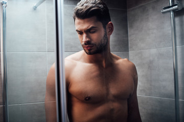 selective focus of serious handsome man standing behind glass shower doors in bathroom