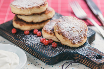 On a white table made of artificial stone is a wooden cutting board with five cottage cheese pancakes and red berries . On the table a red checkered napkin, sour cream, fork and knife.