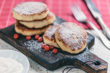 On a white table made of artificial stone is a wooden cutting board with five cottage cheese pancakes and red berries . On the table a red checkered napkin, sour cream, fork and knife.