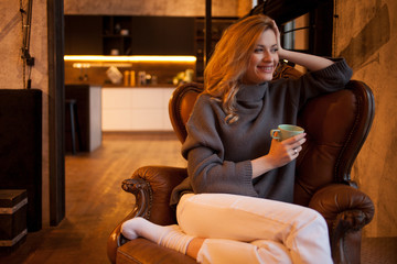 Happy and smiling young blonde is sitting in a cozy chair in her apartment.