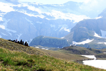 Group of four hikers is taking the rest on the slope and looking on high mountains, snow and frozen lake of Italian Alps