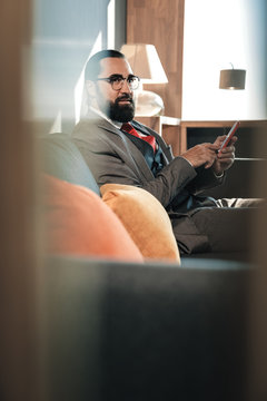 Bearded Businessman Sitting On Grey Sofa With Orange Cushions