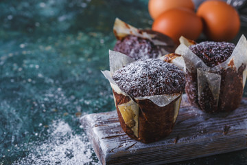 Carrot-chocolate muffin dusted with icing sugar, a cup of tea, baking ingredients. Flour, eggs, lemon citrus on a dark table