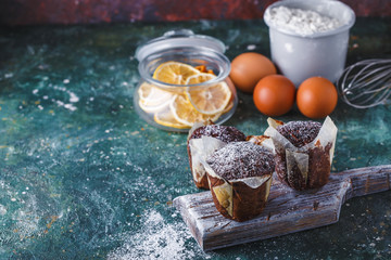 Fototapeta na wymiar Carrot-chocolate muffin dusted with icing sugar, a cup of tea, baking ingredients. Flour, eggs, lemon citrus on a dark table