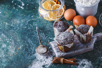 Carrot-chocolate muffin dusted with icing sugar, a cup of tea, baking ingredients. Flour, eggs, lemon citrus on a dark table