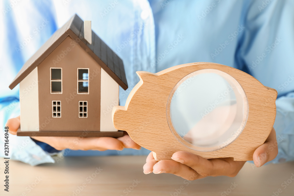 Sticker Woman holding piggy bank with house model at wooden table, closeup