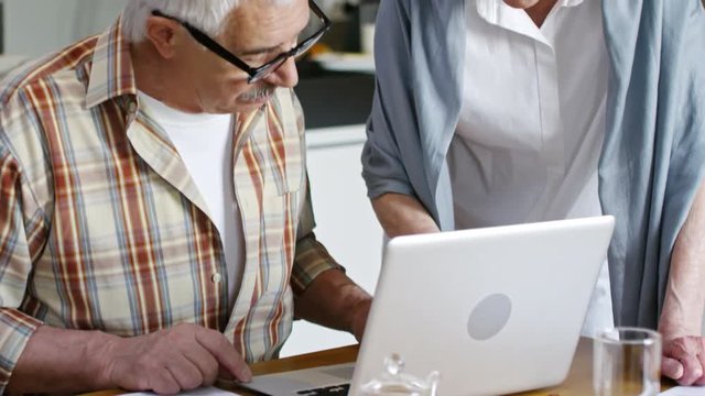 Tilt up of aged woman standing close to aged man who is sitting showing him something on computer
