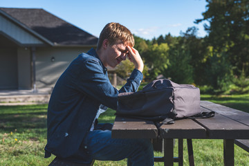 Fototapeta premium Upset teenager sitting at a picnic table in a park all alone. His backpack is lying on the table next to him.
