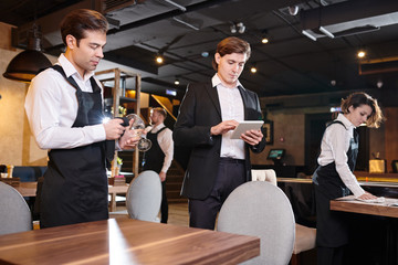 Serious concentrated handsome young restaurant manager using digital tablet while checking online records, waiting staff cleaning tables and crockery