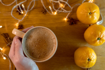 Hand with Cup of coffee  with orange mandarins and cinnamon sticks and anise stars sticks on the wooden table with garland