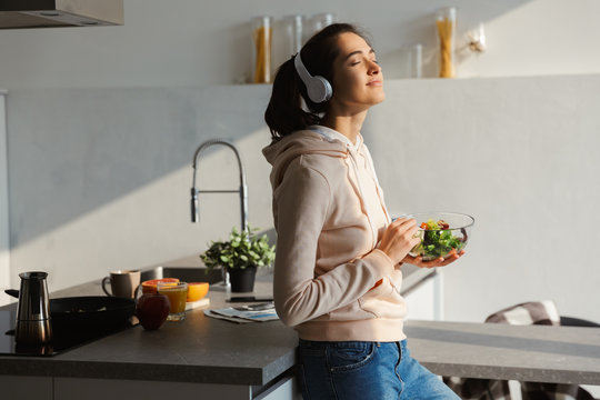 Happy Healthy Woman In The Kitchen Standing Daily Morning Routine Eat Salad Listening Music In Headphones.