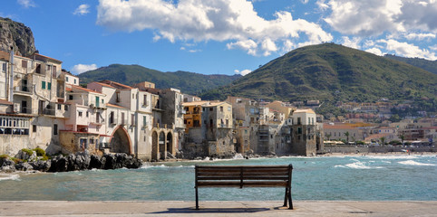 Bench standing on the seafront on the background of the old Sichilian town of Chefalu and green hills on a sunny summer day