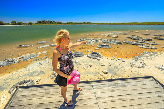 Blonde woman on wooden platform, pointing at Stromatolites on Lake Thetis, a saline coastal lake, Cervantes, Western australia. Tourist enjoys of Australian landscape. Sunny with blue sky.