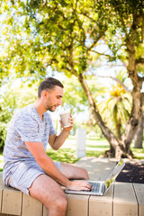 Handsome happy man outdoors in the park using laptop computer drinking coffee.