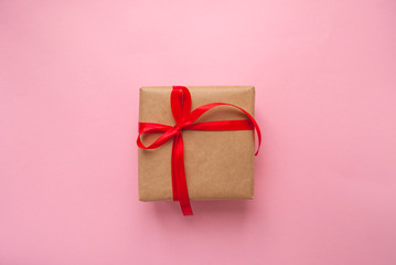 Gift box wrapped in brown colored craft paper and tied with red bow on pink background.