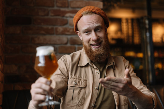 Portrait of happy bearded emotional man holding glass with craft beer, checking quality of alcohol, testing beer and smiling in loft bar. Successful small business concept