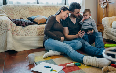 Family with small child and pregnant mother looking at the tablet sitting on the carpet