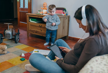 Pregnant woman using the tablet sitting on the carpet in the living room while her son is playing