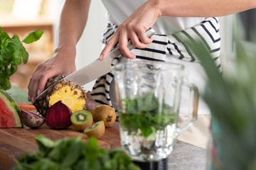 Close-up of a woman's hands cutting a pineapple on a countertop full of fruit and jar of water with mint