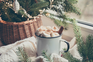 Winter warming mug of chocolate with marshmallow on windowsill with Christmas tree decor.