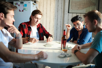Group of young men sitting at the table with bottles of beer dealing cards and betting