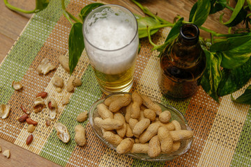 Beer in bottle and glasses and peanuts in crystal bowl. Table with vintage bamboo mat and liana vine in the background.