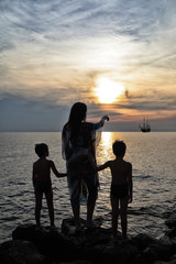 Silhouette - Mother And Her Twins Standing On The Rocks Pointing At Ship On The Horizon On Beautiful Sunset