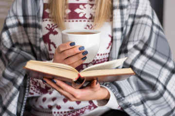 Girl drinking hot tea and reading book in long winter night at home. Woman warming and covered with retro blanket. Cold weather and winter concept. Close up, selective focus