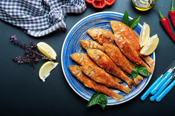 Fried seafood (‘red mullet’, ‘barbule’) on on a blue plate, dark background. Next to the slices of lemon and basil flowers
