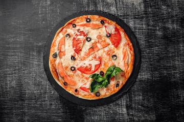 Pizza with cheese, trout, tomatoes, olives and shrimps on a stone and a black chalk board, top view.