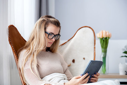 Young woman in glasses reading ebook using digital reader while sitting in comfortable armchair at home
