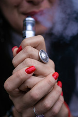 Caucasian woman with red nails manicure and antique ring on finger holds small vape. Smoking alternative vay. Woman exhales thick smoke. Life without cigarettes. Woman-vaper. Small e-cigarette.