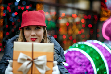 Fototapeta na wymiar Positive blonde woman with red lipstick wearing trendy cap and holding gift box with a bow. Space for text