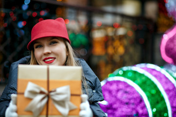 Fototapeta na wymiar Happy blonde woman with red lipstick wearing trendy cap and holding gift box with a bow. Space for text