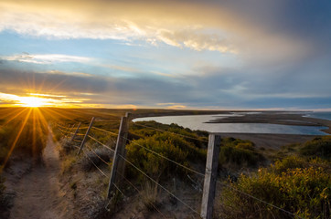 Fototapeta premium Landscape with a lake and a road with a wire fence on sunset with a golden and blue sky and sun rays