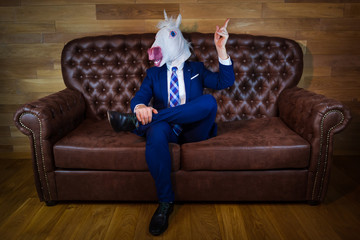 Funny unicorn in elegant suit siting on sofa like a boss and showing hand gesture with raised index finger. Unusual man at home. Freaky guy in mask.