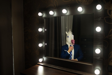 Glamour man in elegant suit and funny mask corrects tie and looks at himself in mirror in dressing...