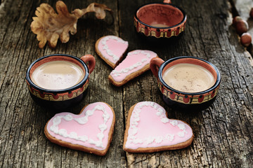 Two full cups of coffee next to two heart-shaped gingerbreads are close to each other and one empty coffee cup next to a broken heart-shaped gingerbread at a distance on old wooden boards. 