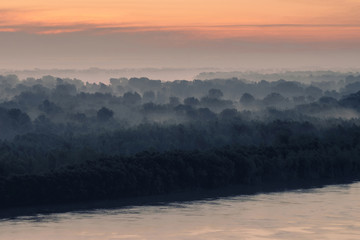 Mystical view on riverbank of large island with forest under haze at early morning. Eerie mist among layers from tree silhouettes. Morning atmospheric landscape of majestic nature in blue faded tones.