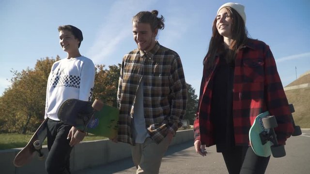 A company of smiling young people carry skateboards in their hands while walking in the park. Skateboarders spend time outdoors. Hobbies and lifestyle.